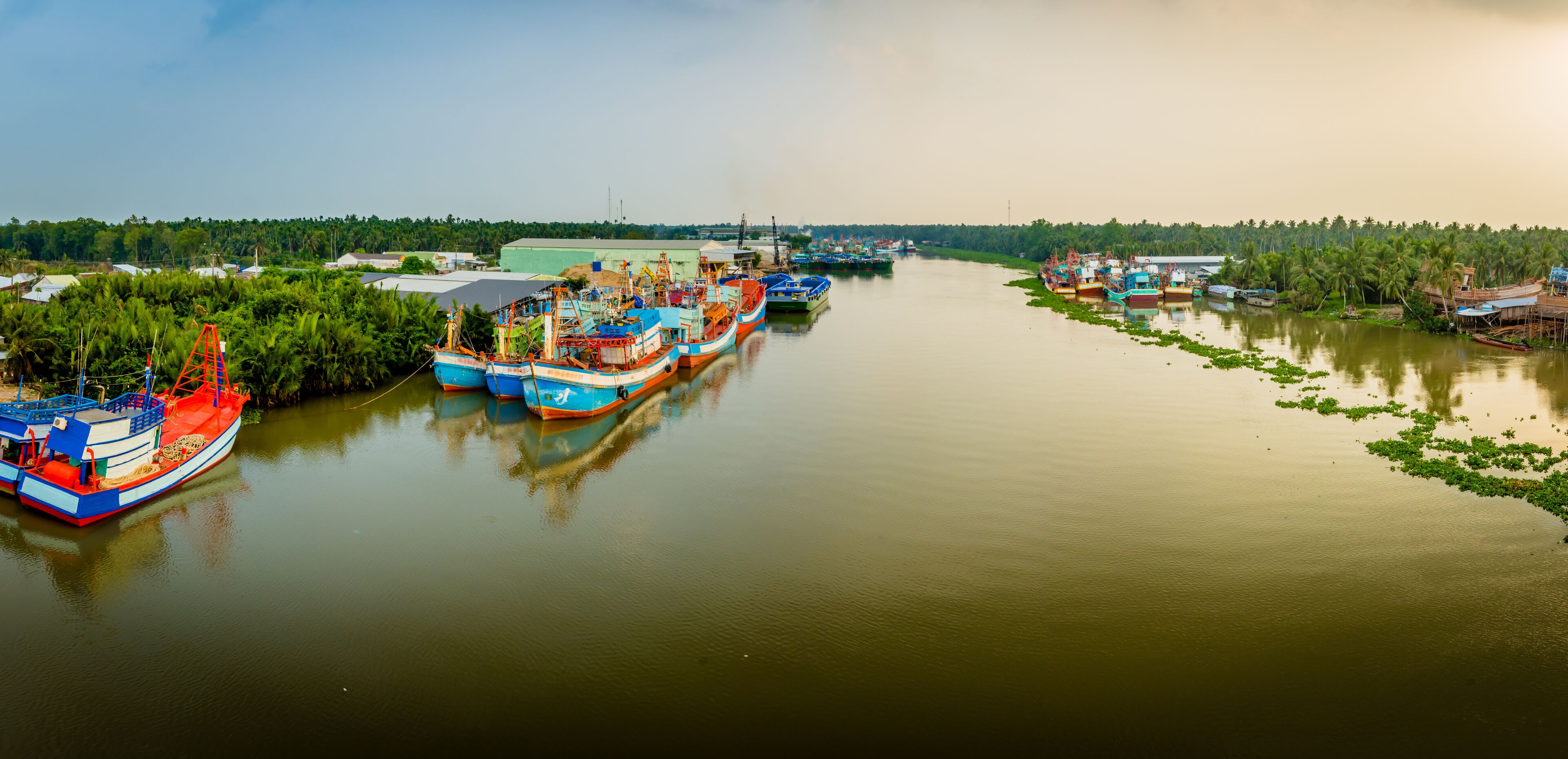 A river with several boats docked along the banks, surrounded by greenery and trees.