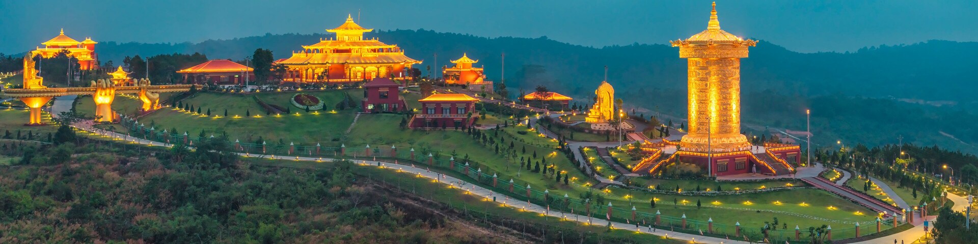 Un complejo de templos bien iluminado con múltiples pagodas y una torre alta, rodeado de exuberante vegetación y colinas al fondo.