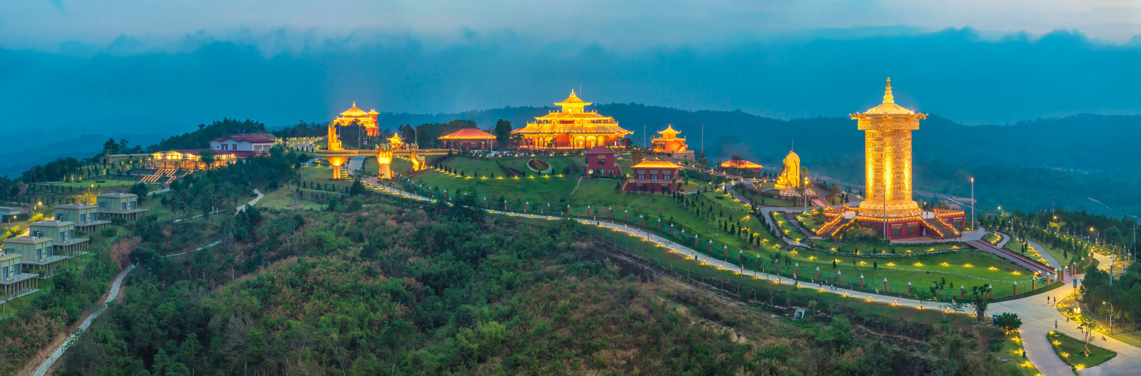 A well-lit temple complex with traditional architecture, surrounded by lush greenery and hills.