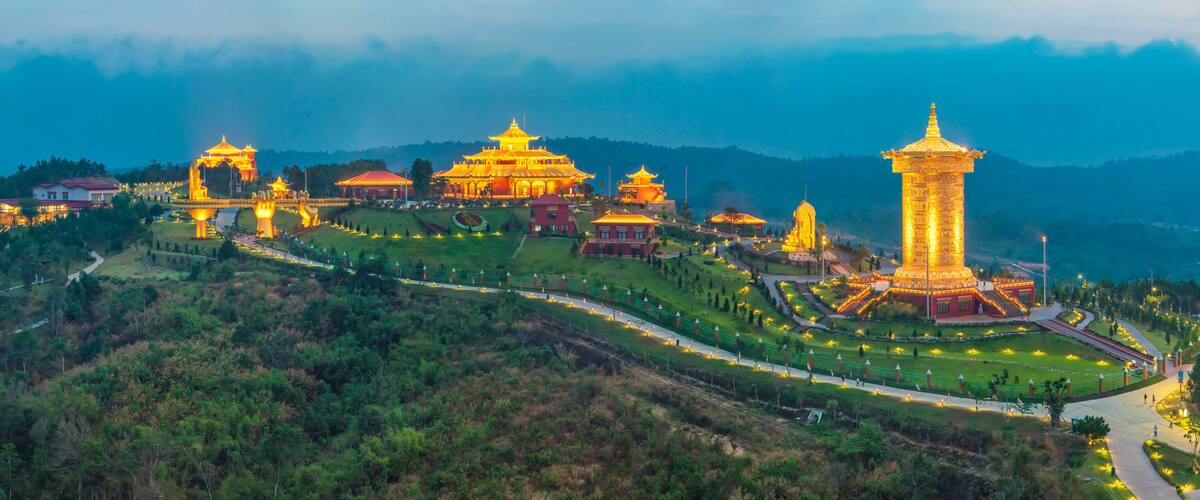 A well-lit temple complex with traditional architecture, surrounded by lush greenery and hills.
