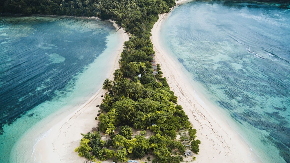 Eine Vogelperspektive zeigt eine schmale Insel mit dichtem Grün, umgeben von türkisblauem Wasser und einem klaren Himmel.