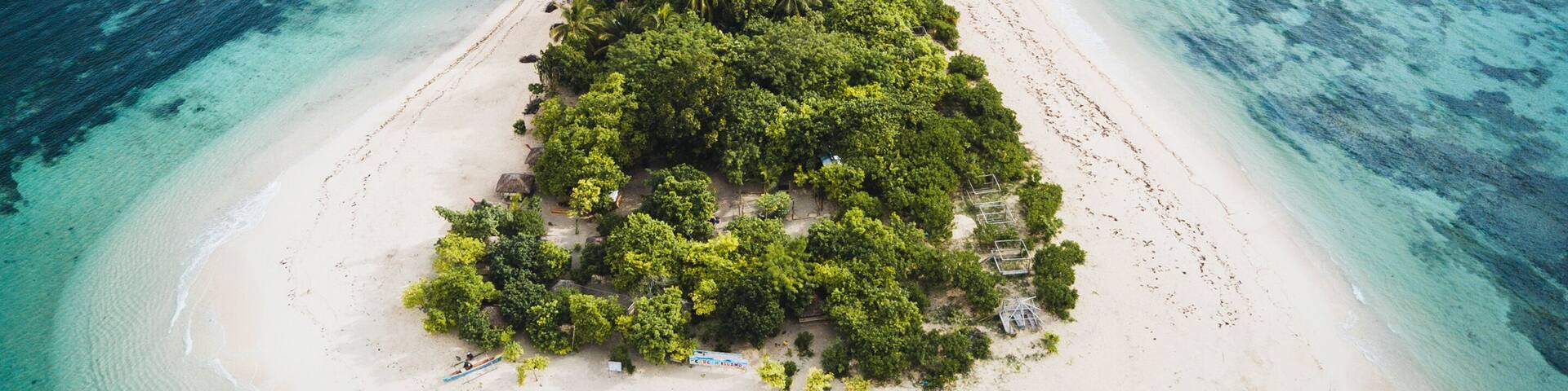 An aerial view of a narrow island with dense green vegetation, surrounded by turquoise waters and a clear sky.