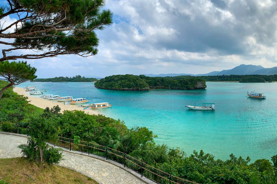 A coastal landscape with turquoise waters, boats, a sandy beach, and lush greenery.