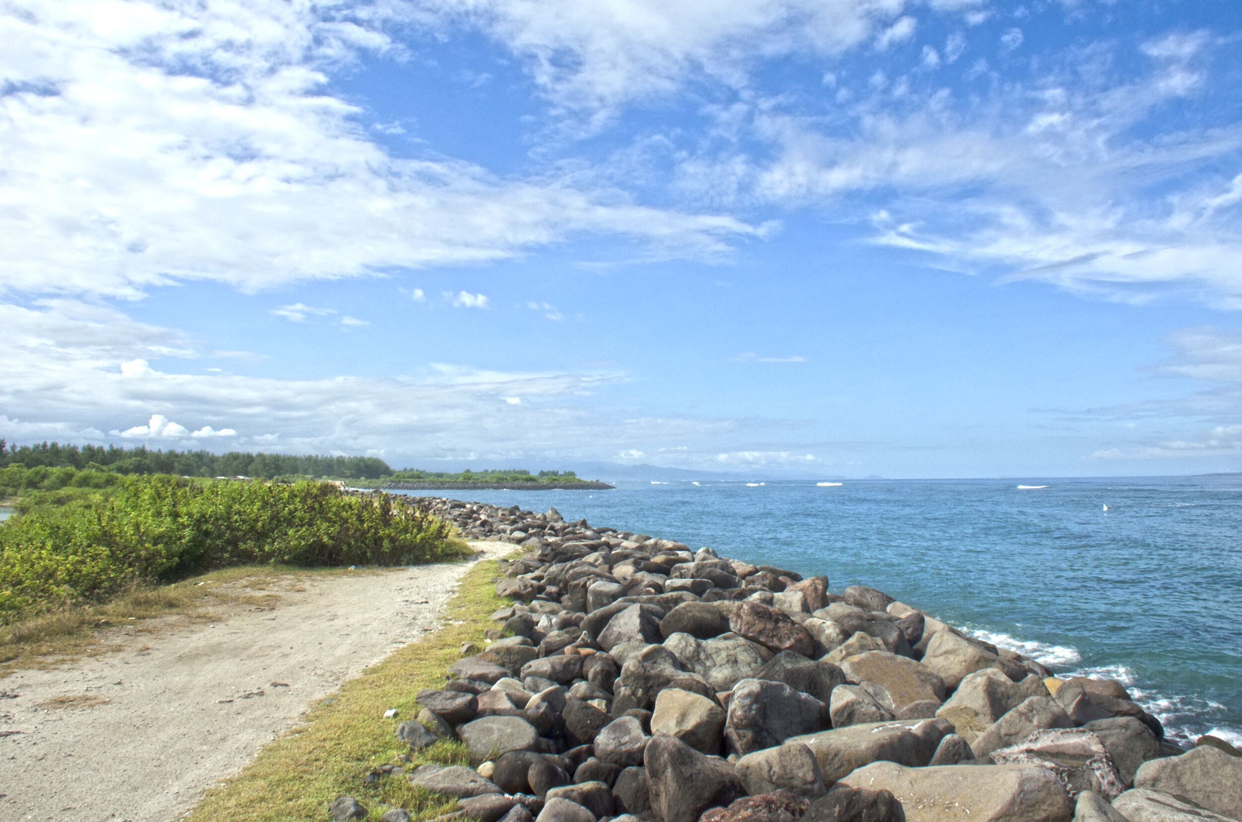 A rocky shoreline with a dirt path leading to the water, surrounded by greenery and a clear blue sky.