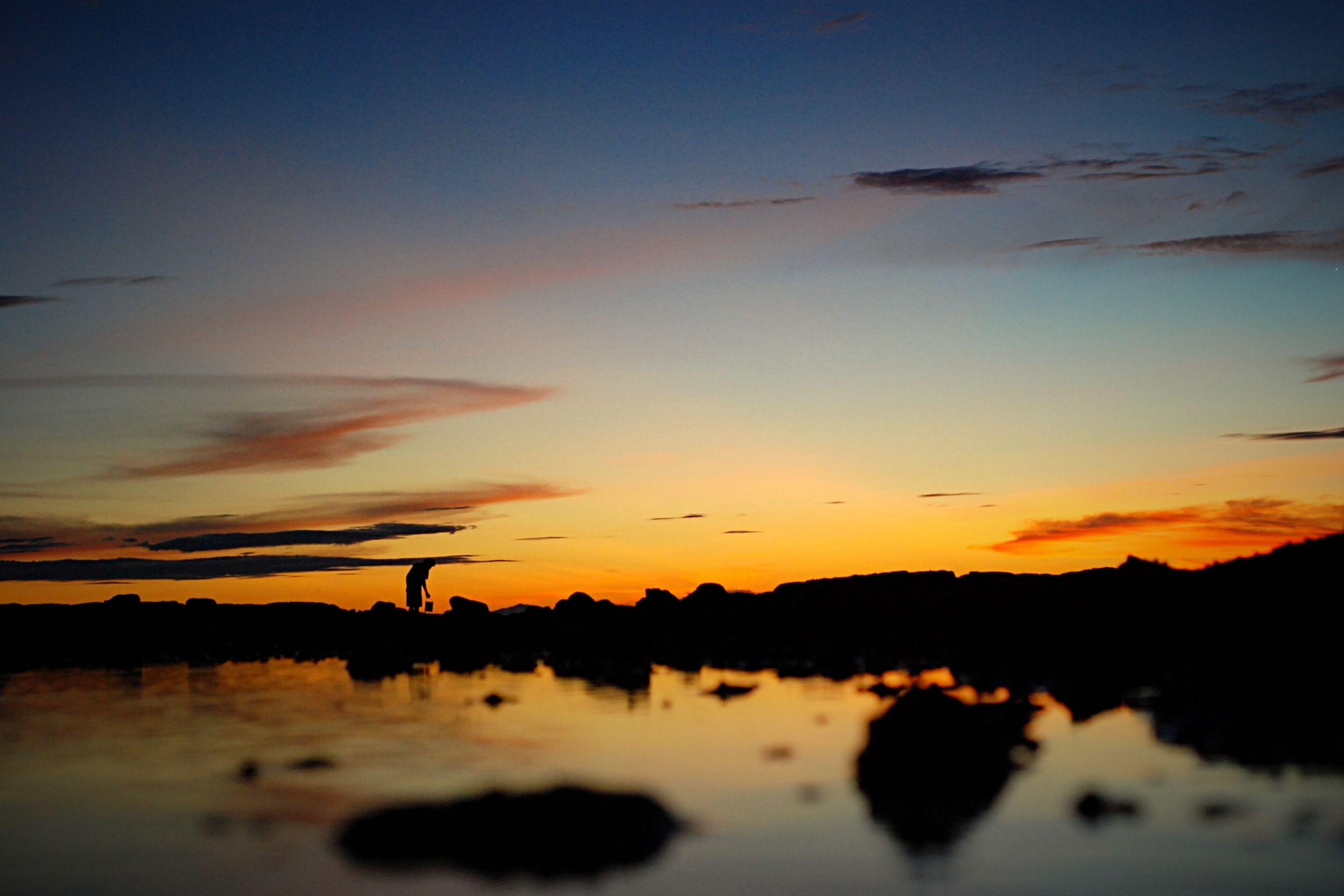 A sunset over a body of water with silhouettes of rocks and a person standing on the shore.