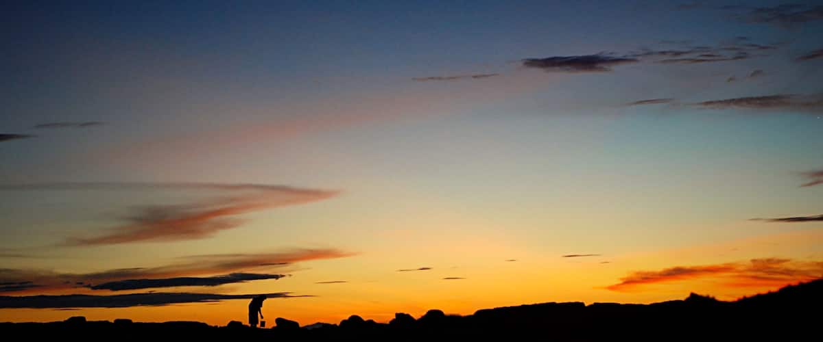 A sunset over a body of water with silhouettes of rocks and a person standing on the shore.