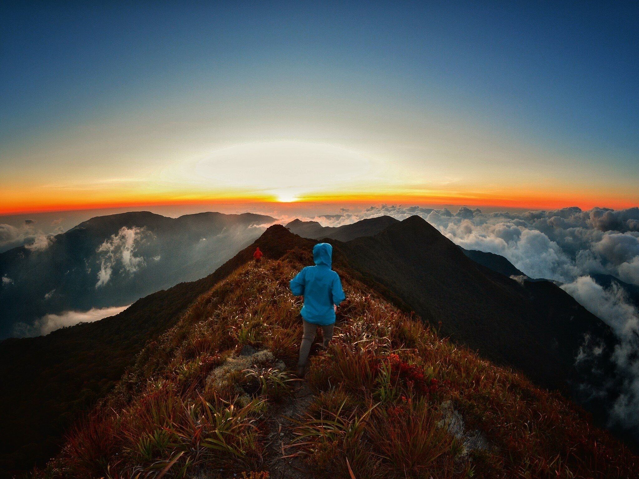 A person in a blue jacket walking on a mountain ridge with a sunset in the background.