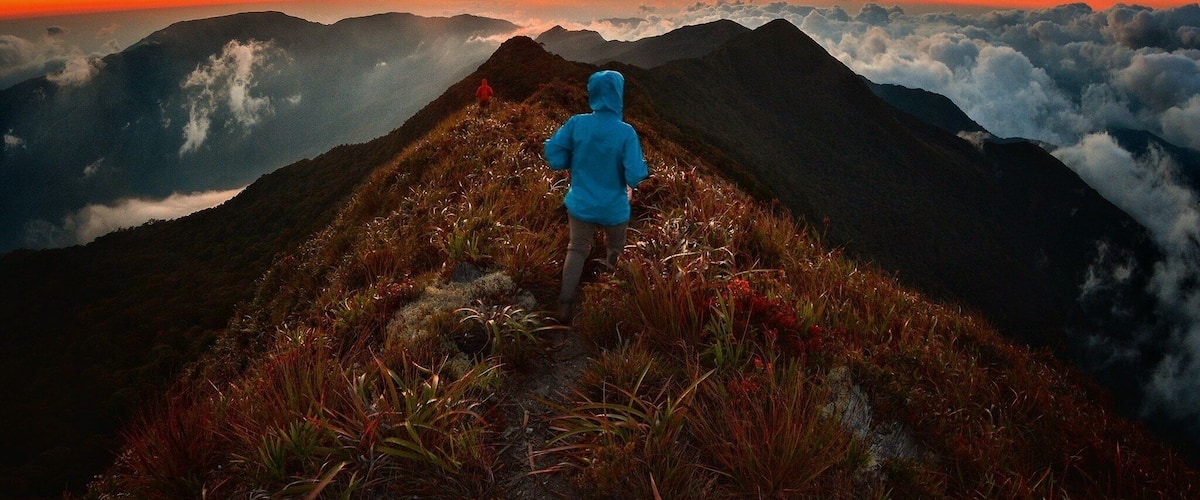 A person in a blue jacket walking on a mountain ridge with a sunset in the background.