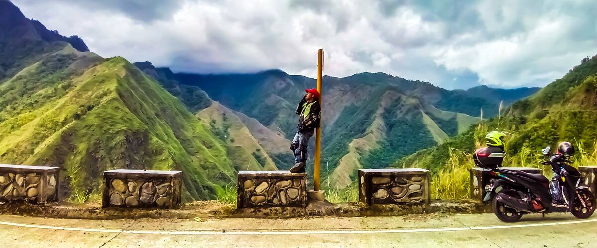 A person standing on a stone ledge with a mountain range in the background.
