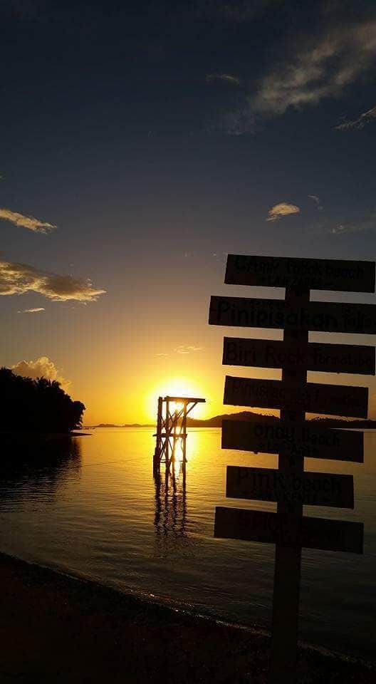 A sunset over a calm lake with a pier and a signpost with multiple destinations.