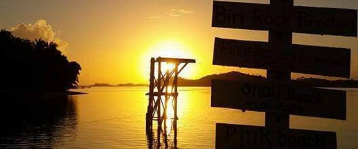 A sunset over a calm lake with a pier and a signpost with multiple destinations.