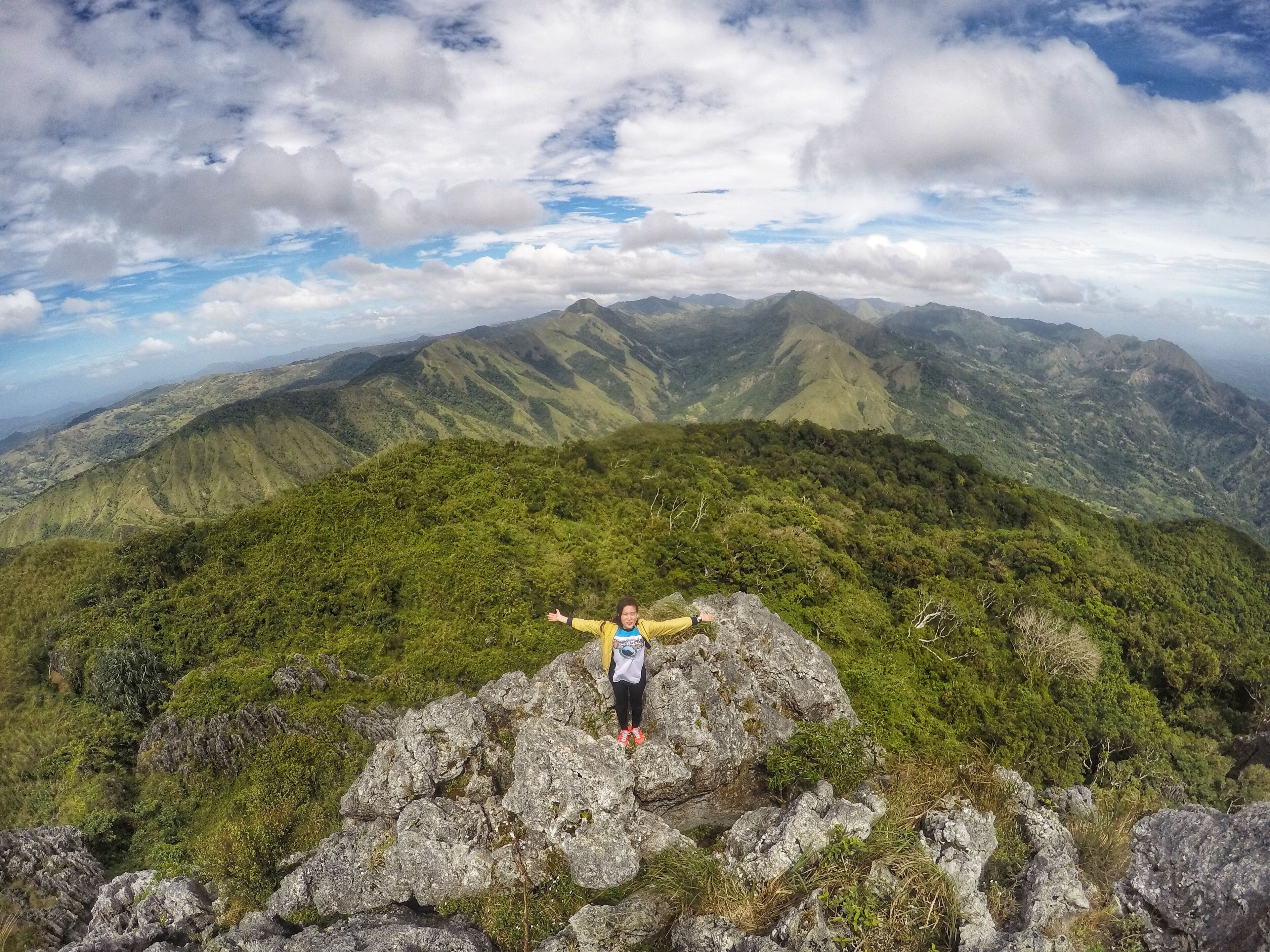 A person standing on a rocky outcrop with arms outstretched, overlooking a mountainous landscape with lush greenery and distant peaks under a partly cloudy sky.