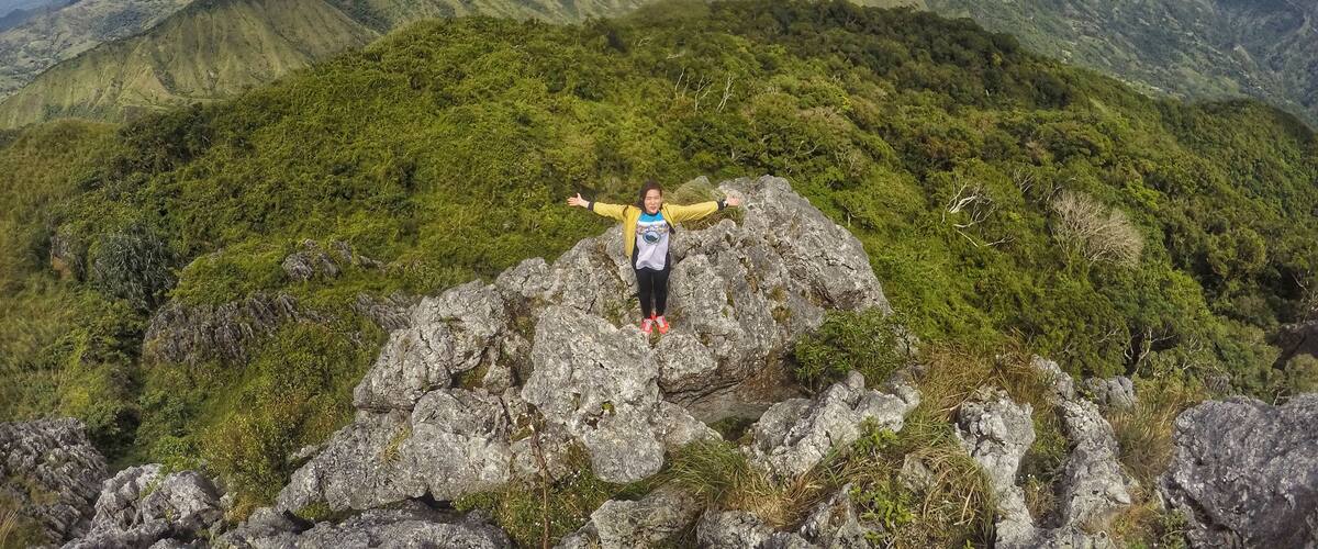 A person standing on a rocky outcrop with arms outstretched, overlooking a mountainous landscape with lush greenery and distant peaks under a partly cloudy sky.
