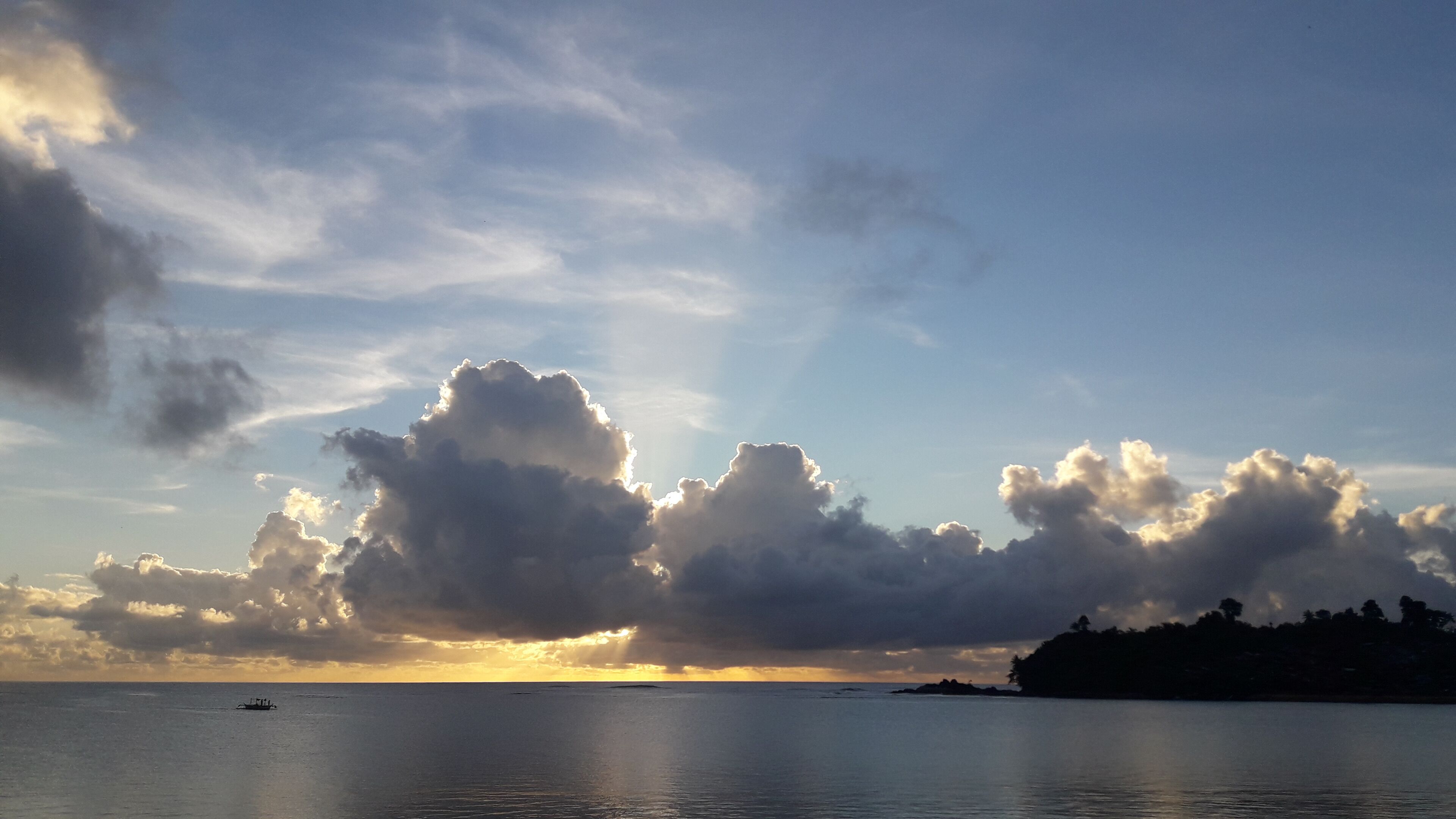 A sunset over a calm sea with large clouds and a distant shoreline.