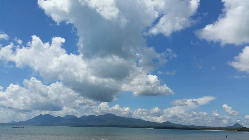 A clear sky with scattered clouds over a calm sea and distant mountains.