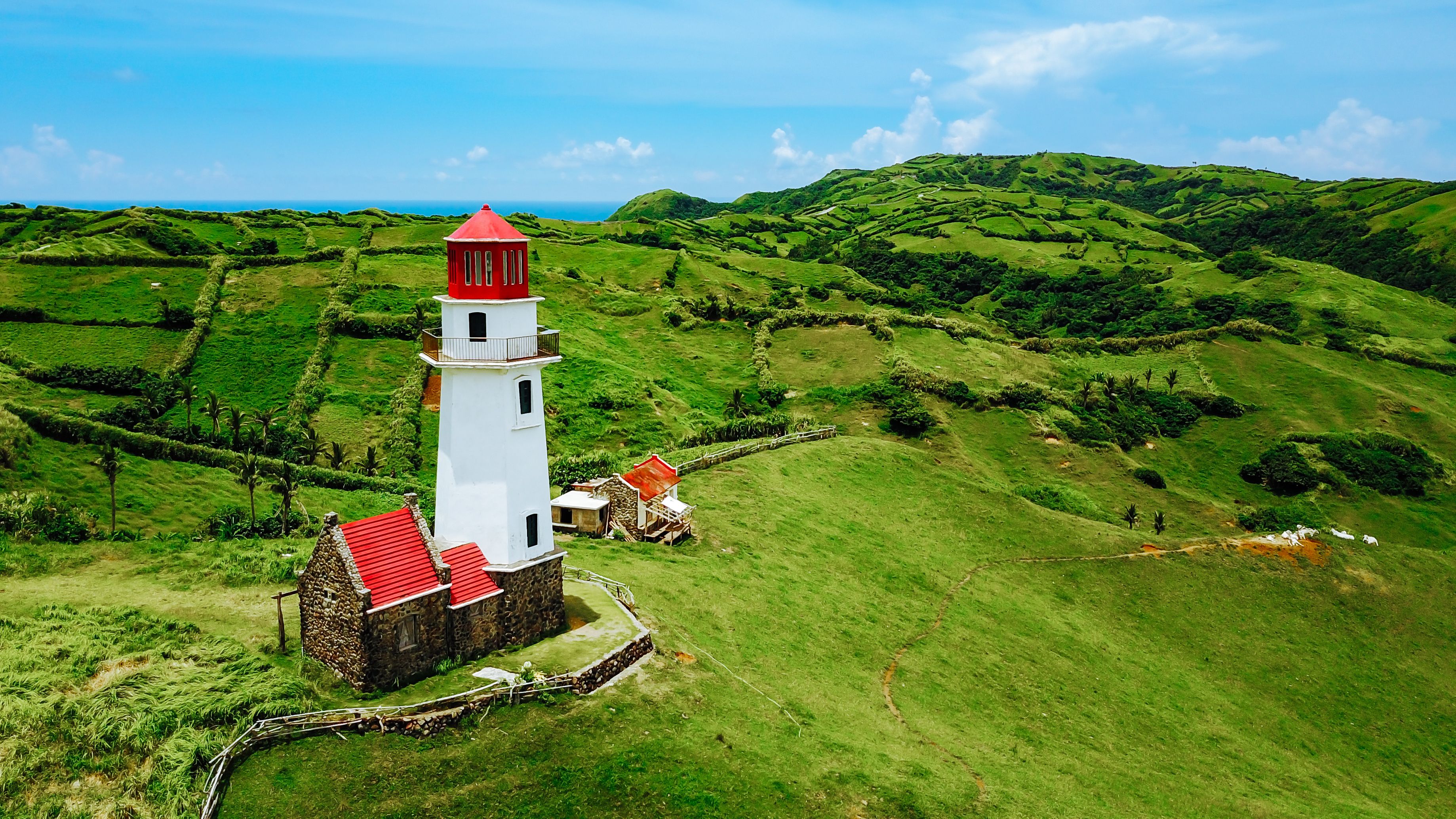 A lighthouse on a grassy hill with a red roof, surrounded by lush green fields and a clear blue sky.