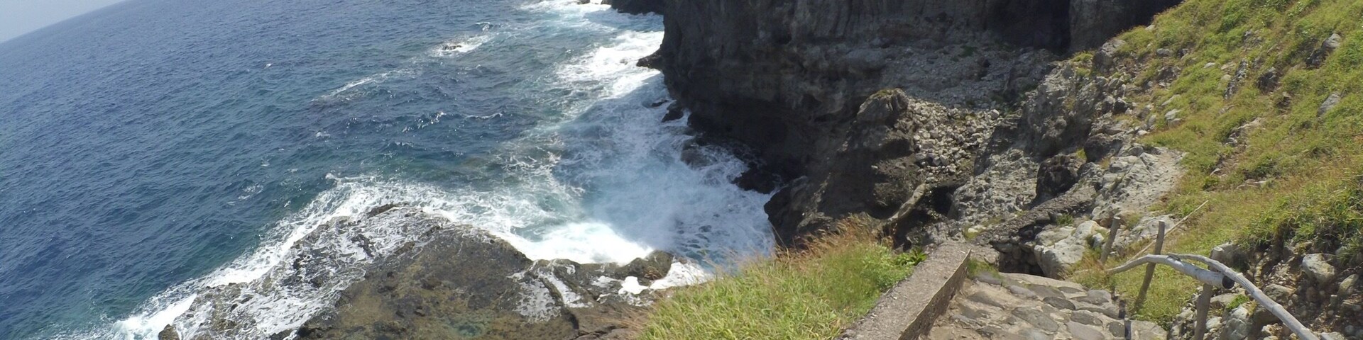 Acantilado costero con olas rompiendo, zona con césped y una escalera de piedra.