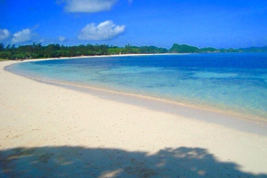 Una playa arenosa con aguas azules cristalinas y colinas verdes al fondo.