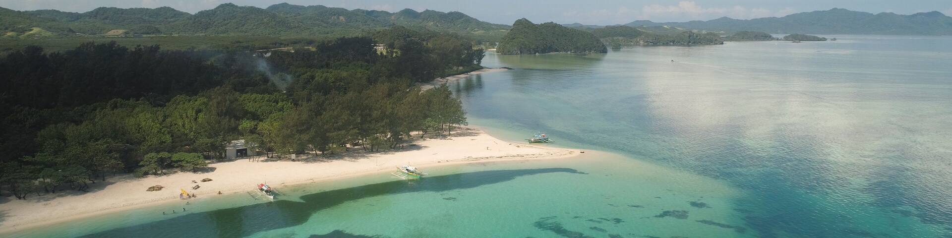 A coastal landscape with a sandy beach, clear turquoise waters, and a backdrop of green hills.