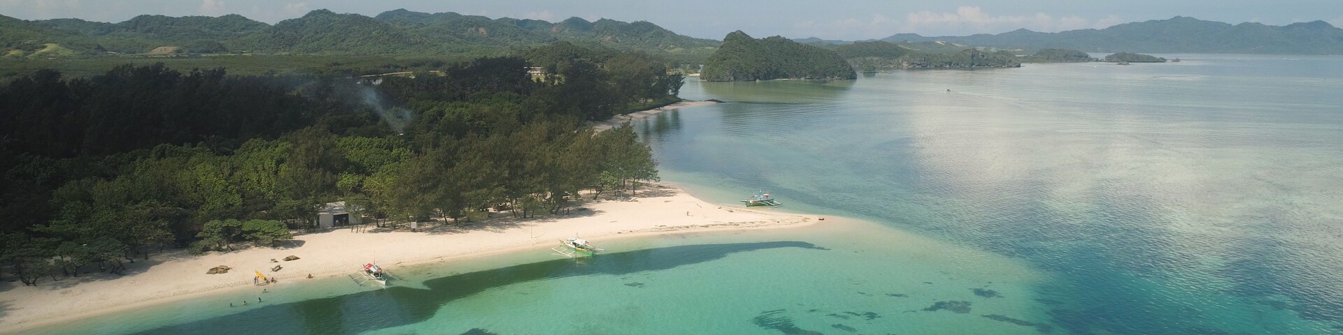 A coastal landscape with a sandy beach, clear turquoise waters, and a backdrop of green hills.
