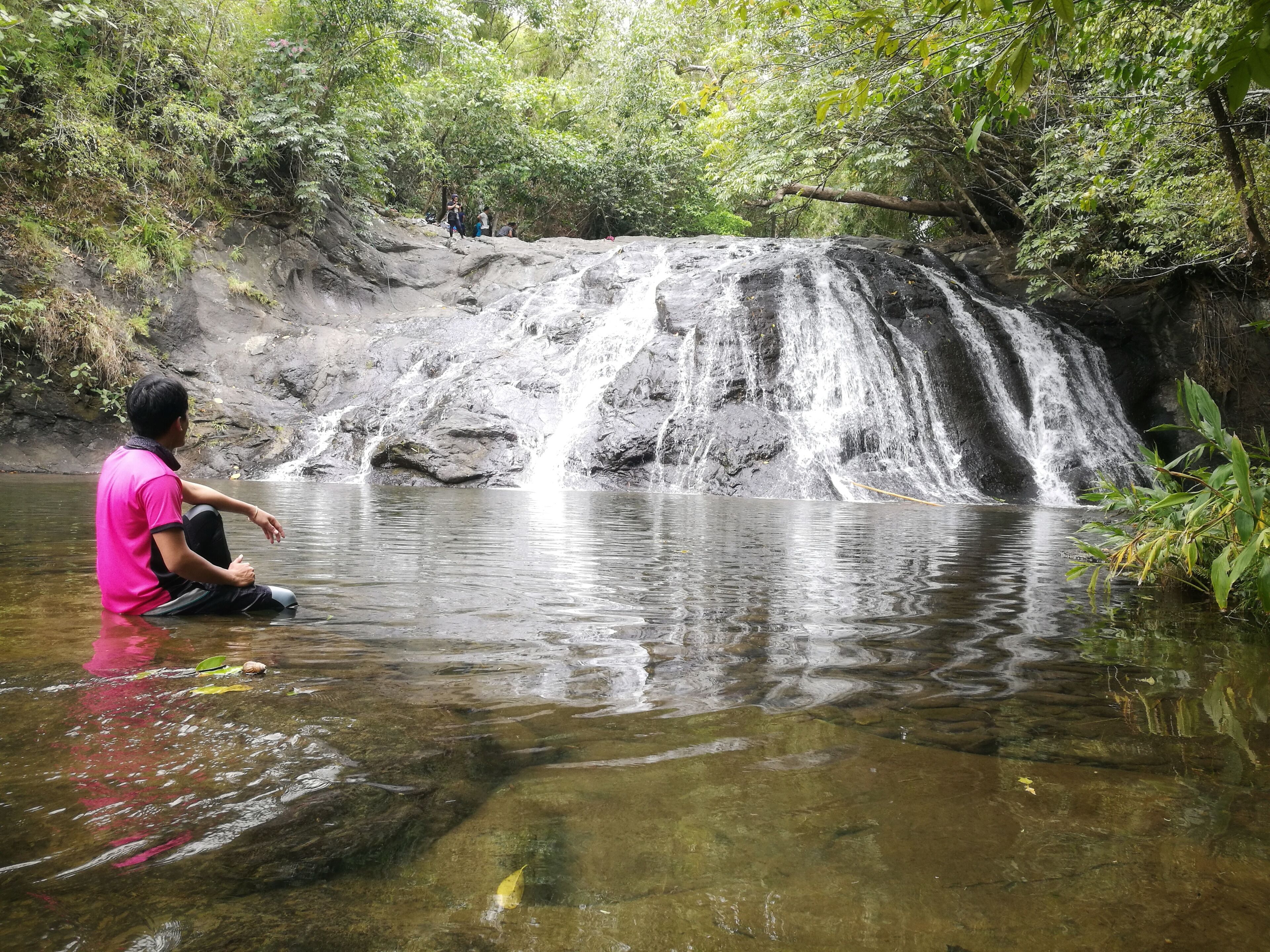 A waterfall in a forested area with clear water and a person sitting by the water.