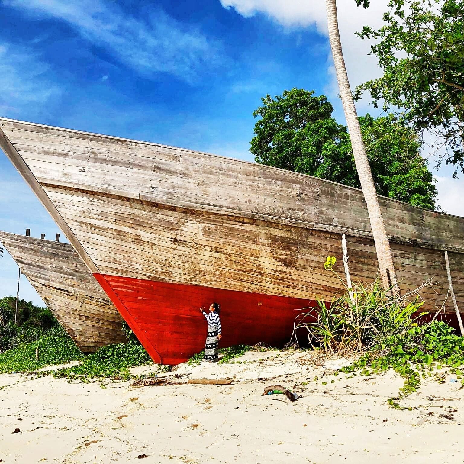 A wooden boat with a red hull resting on a sandy beach, surrounded by greenery and palm trees.