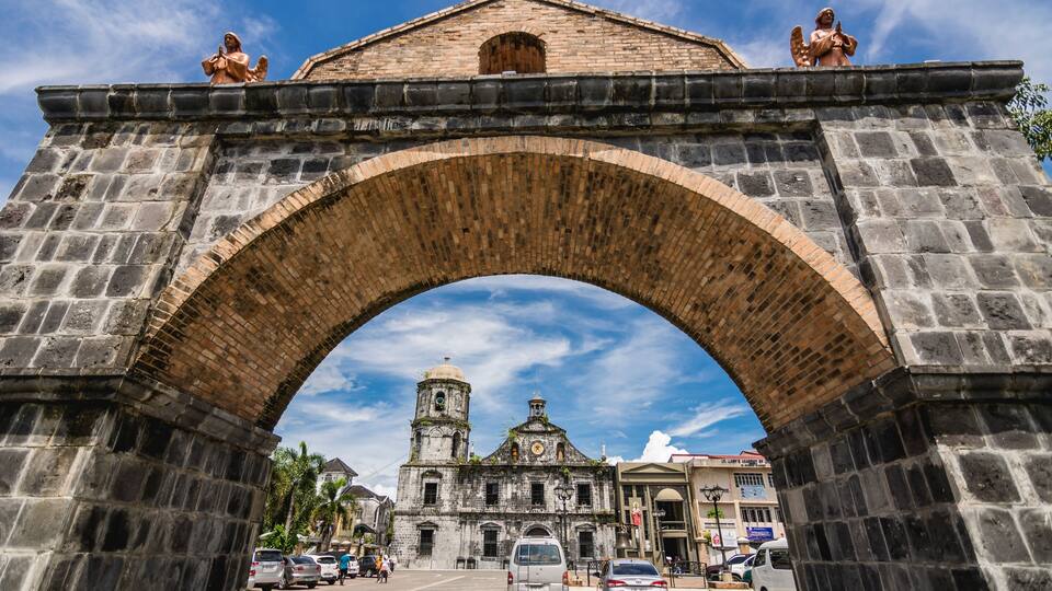 A stone archway with statues, leading to a historic building with a bell tower.