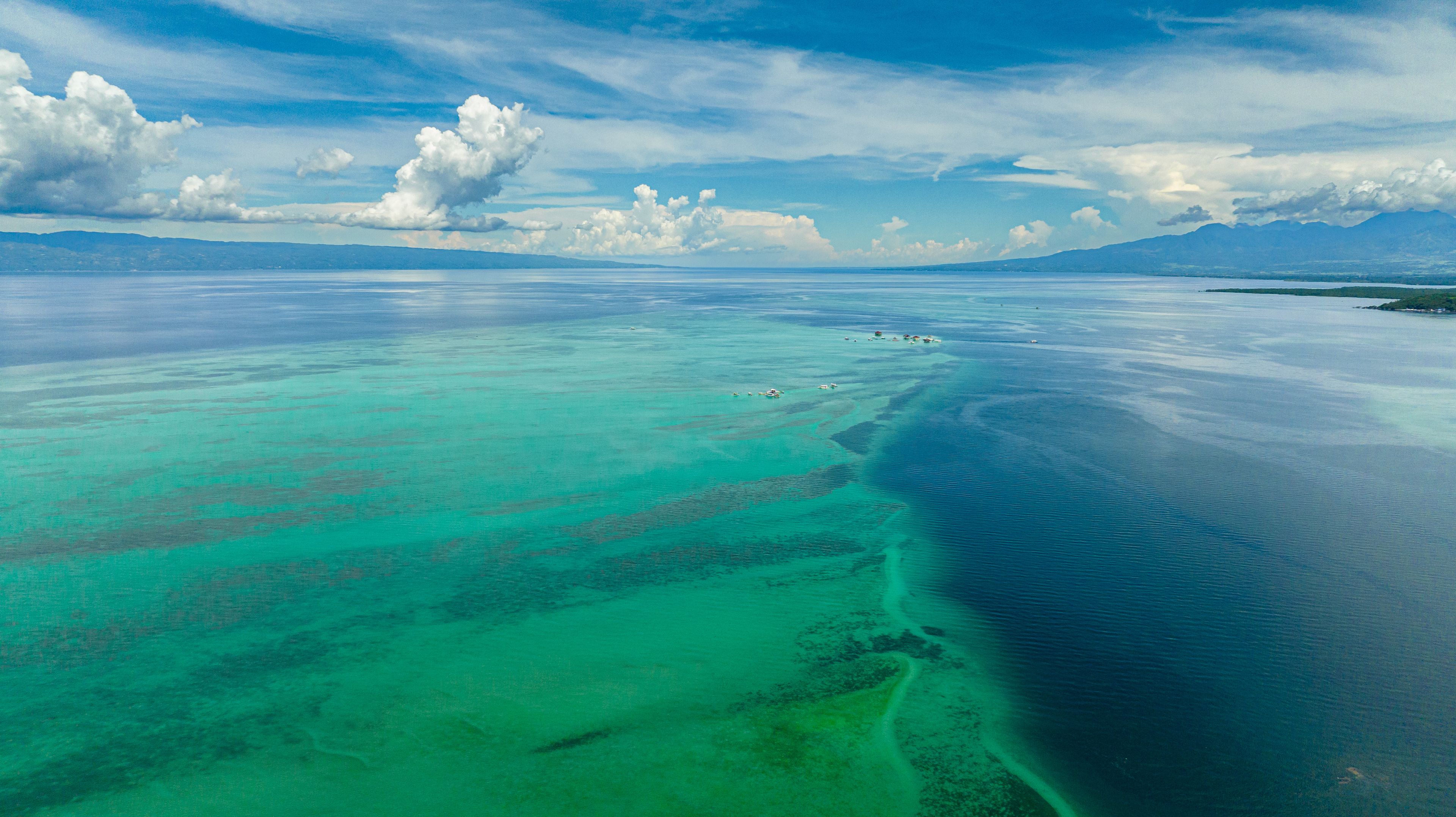 A vast turquoise sea with clear demarcations of shallow and deep waters, a distant shoreline, and a range of mountains under a partly cloudy sky.