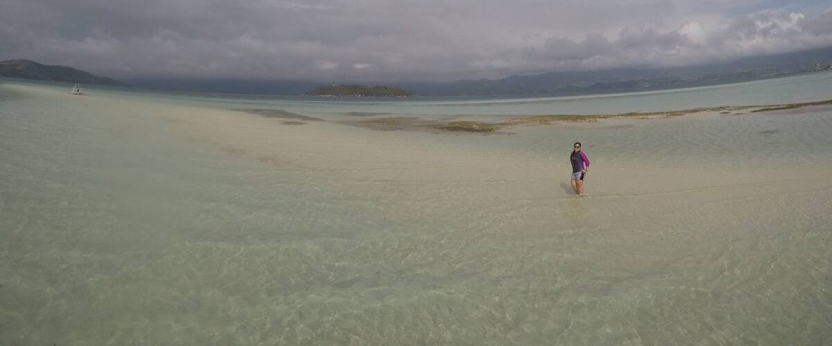 A person standing on a beach with clear water and distant islands under a cloudy sky.