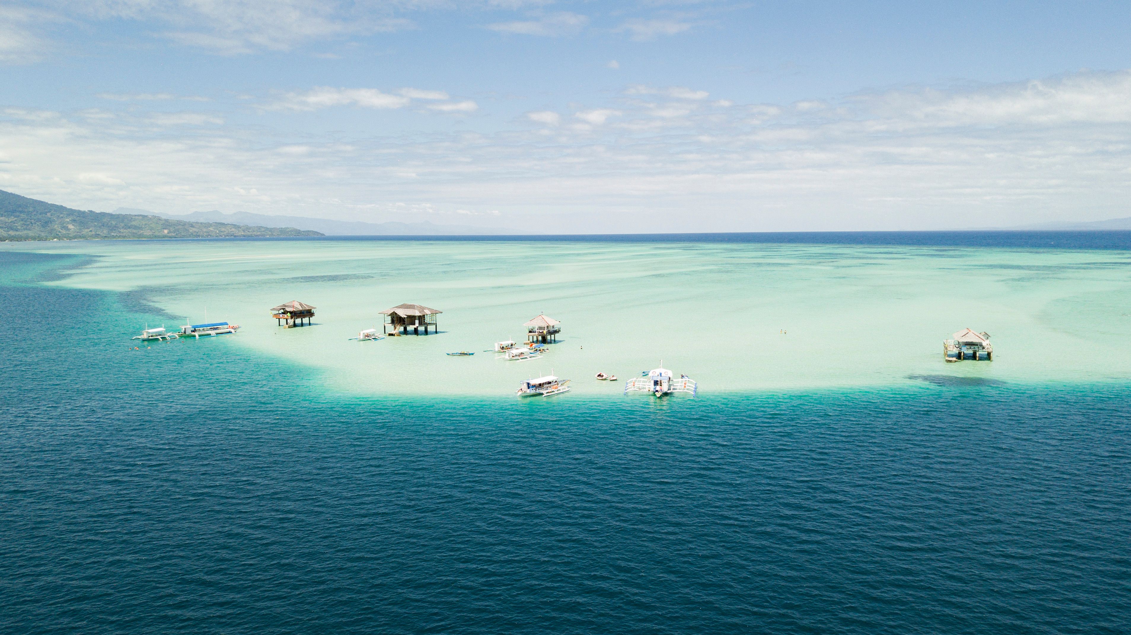 A tropical island with clear turquoise waters, several overwater bungalows, and a few boats.