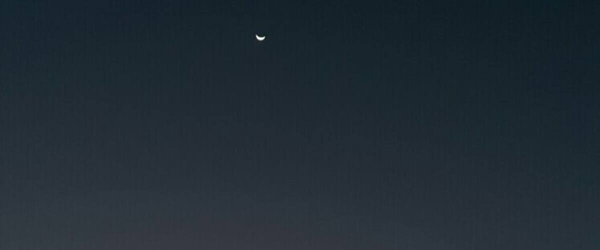A sunset over a calm sea with a crescent moon in the sky.