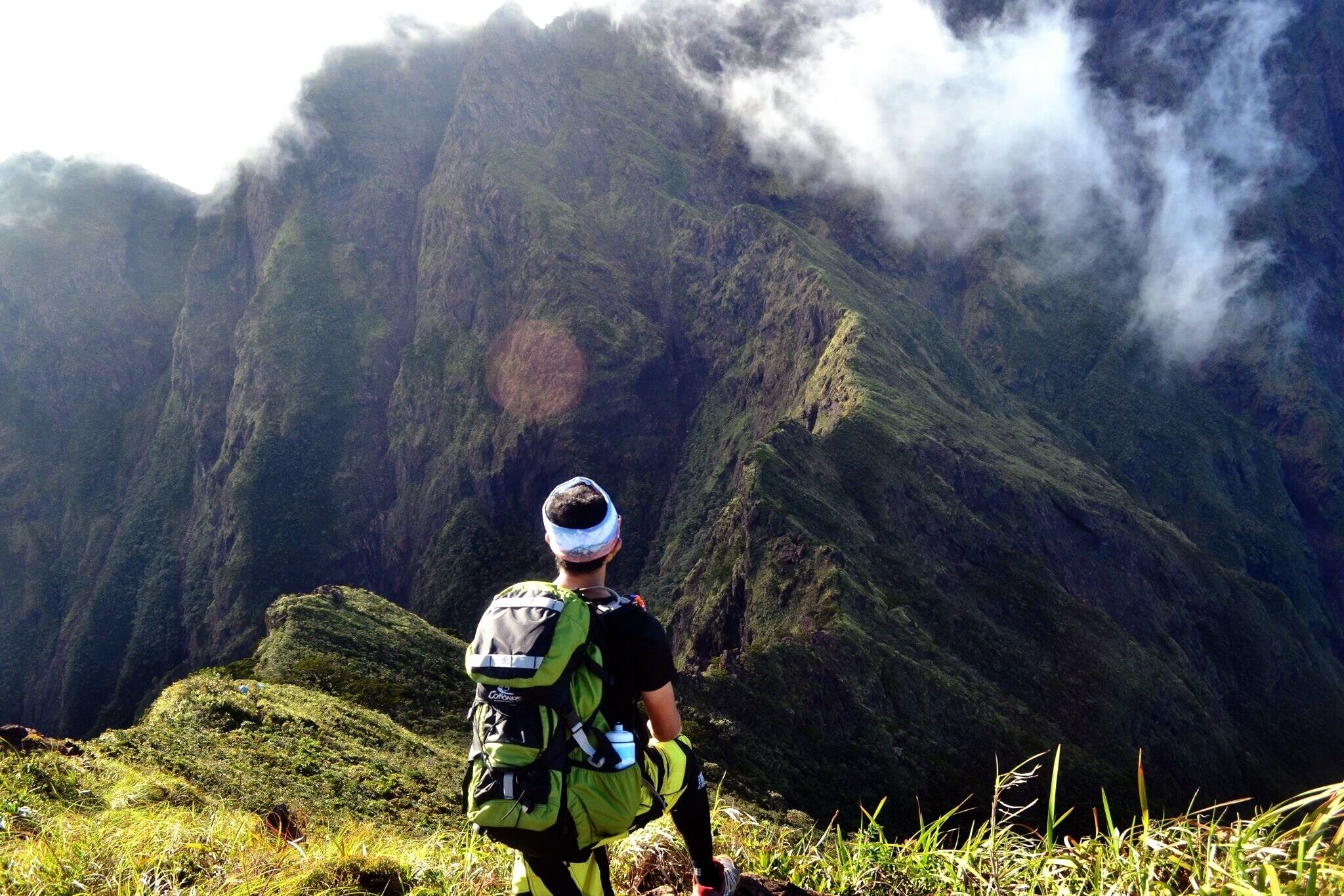 A hiker with a green backpack and a headlamp, overlooking a steep, green mountain with a visible cloud formation.