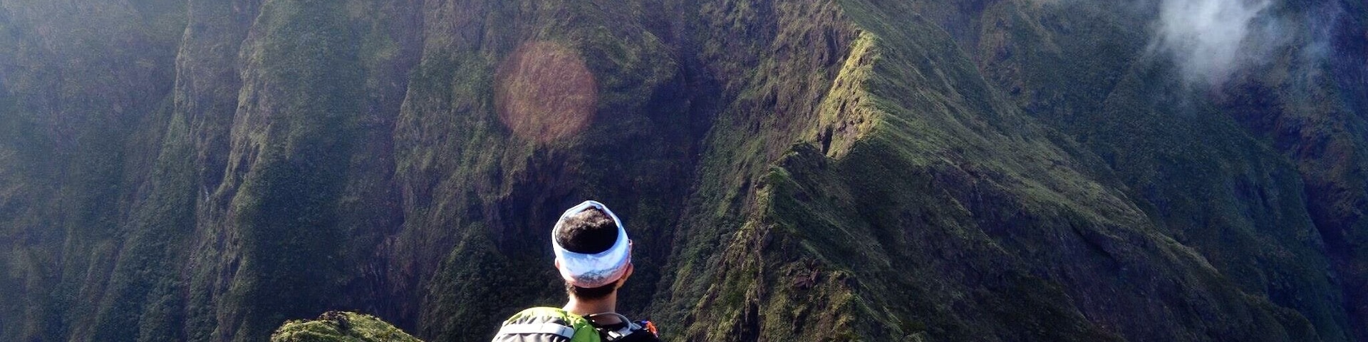 A hiker with a green backpack and a headlamp, overlooking a steep, green mountain with a visible cloud formation.