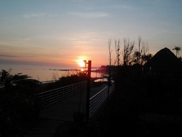 A sunset over a beach with a wooden walkway and a thatched-roof structure.