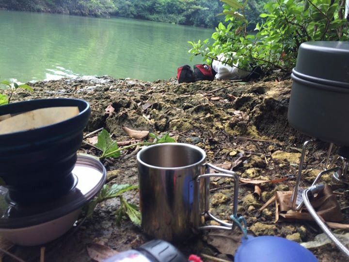 A camping setup near a lake with a metal mug, a bowl, and a pot on a portable stove.