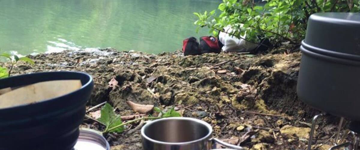 A camping setup near a lake with a metal mug, a bowl, and a pot on a portable stove.