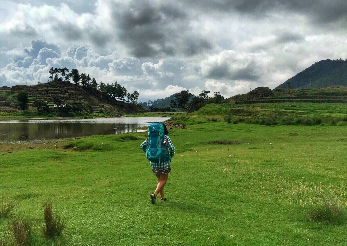 A person walking on a grassy field with a lake and hills in the background.