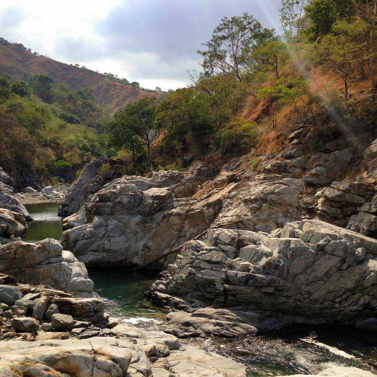 A rocky riverbed with a small stream, surrounded by hills and trees.