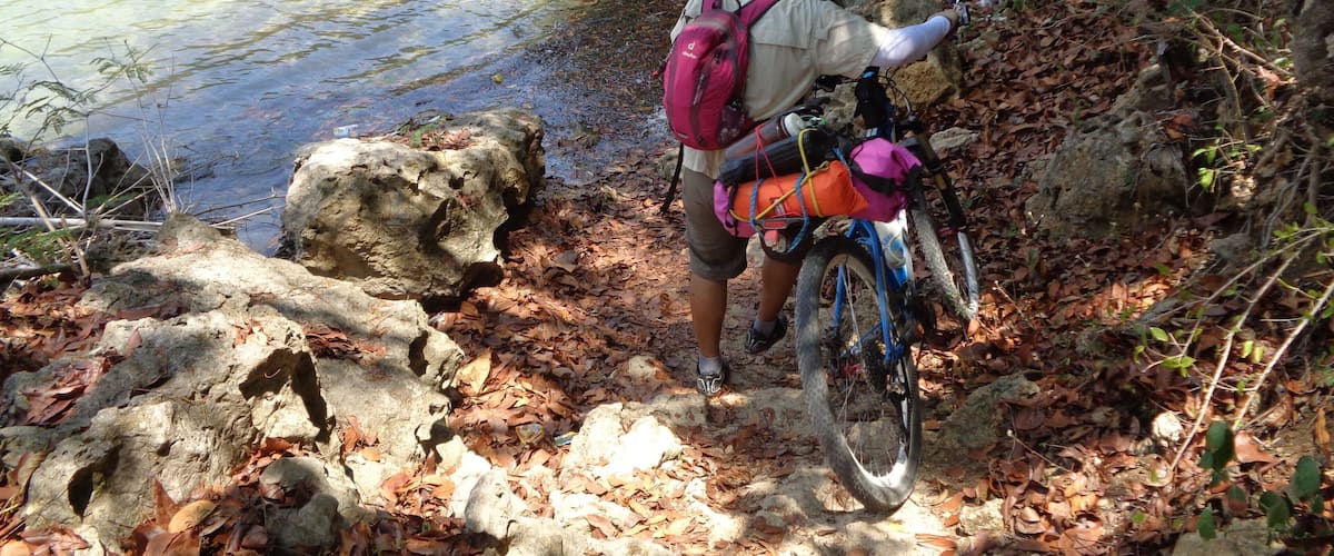 A person with a bicycle and a backpack near a rocky shore by a clear lake.