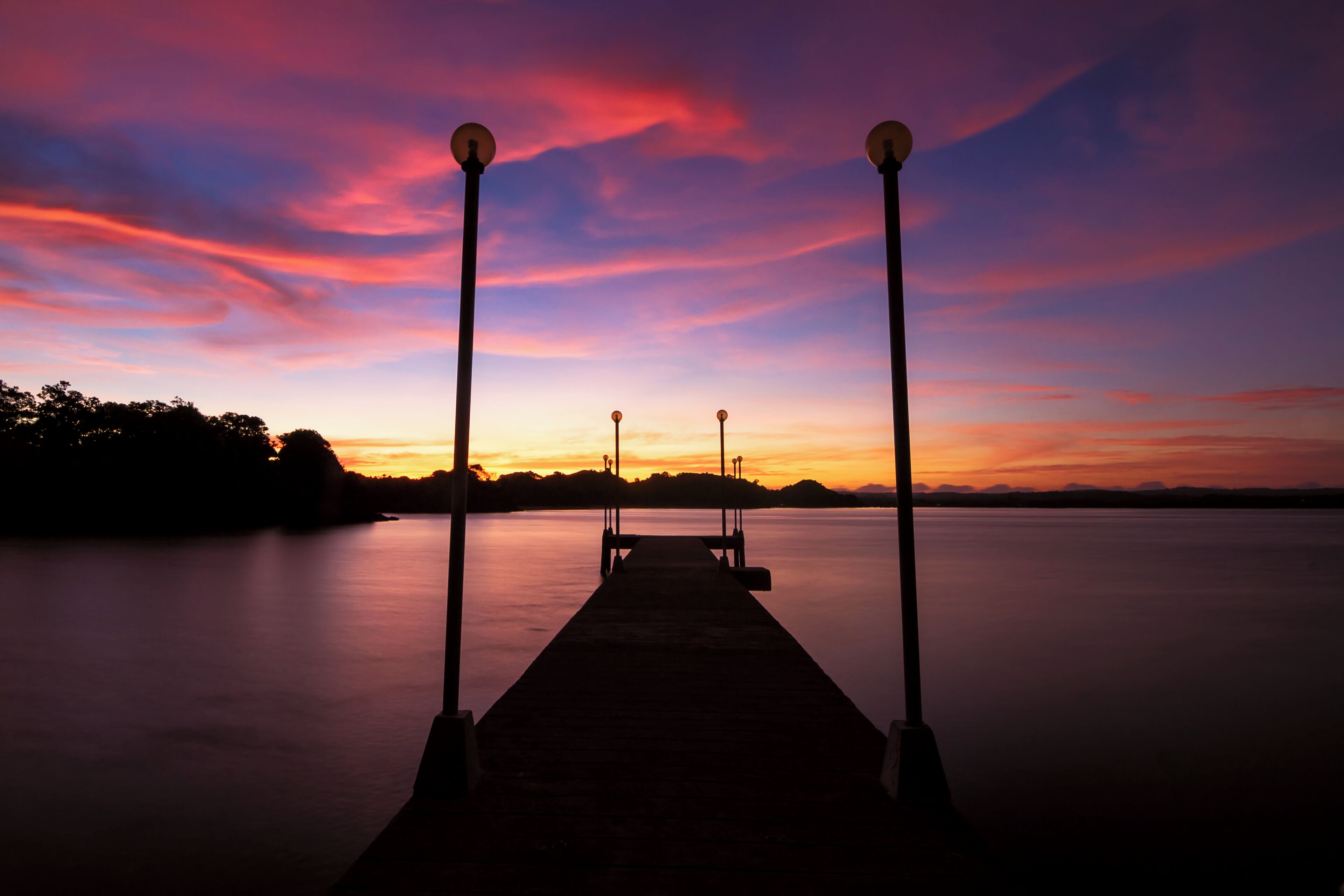 A sunset over a calm lake with a wooden dock and street lamps.