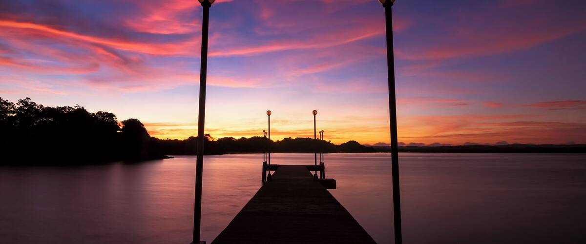 A sunset over a calm lake with a wooden dock and street lamps.