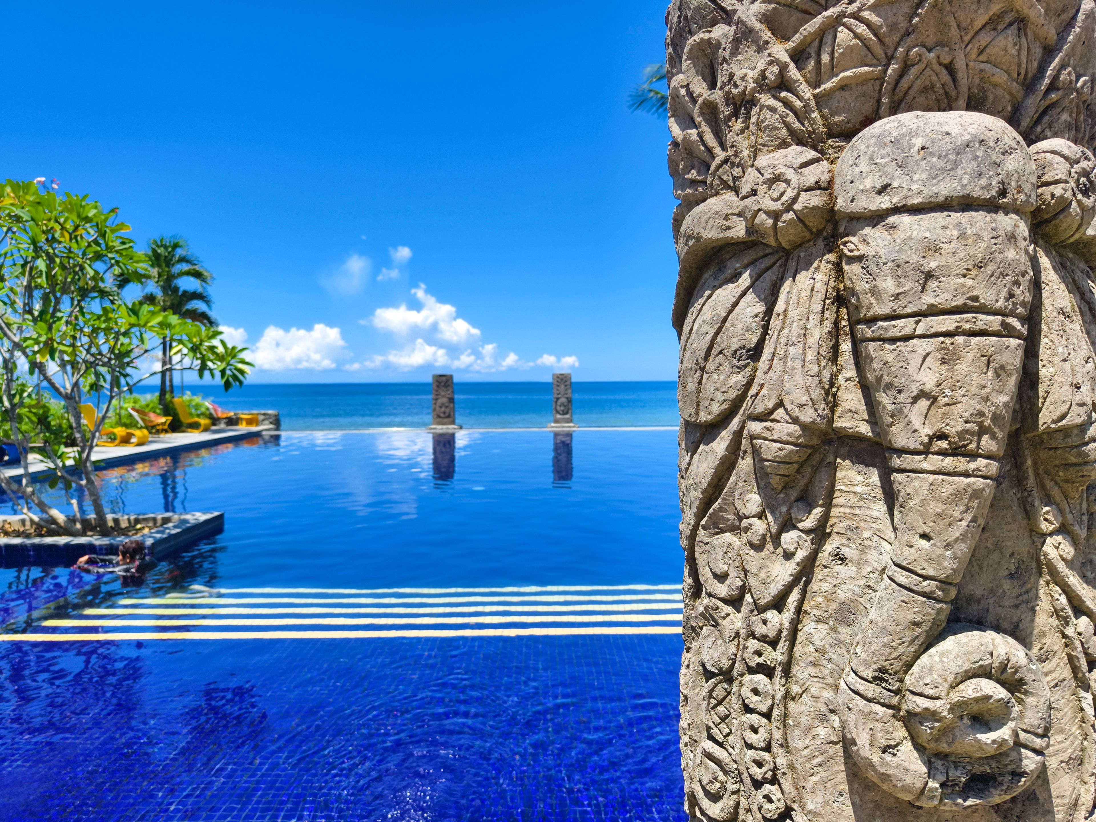 A clear blue pool with a stone sculpture, palm trees, and a view of the ocean.