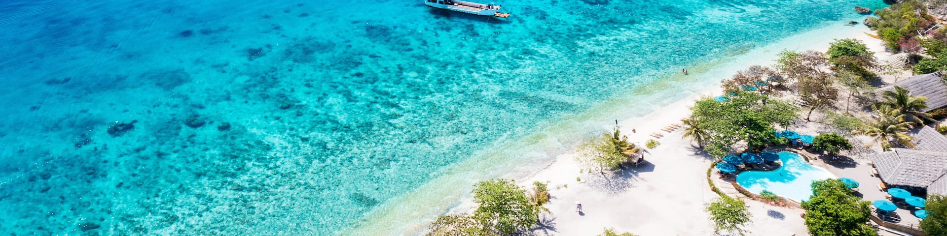A turquoise sea with boats, a sandy beach, and a pool area with umbrellas.