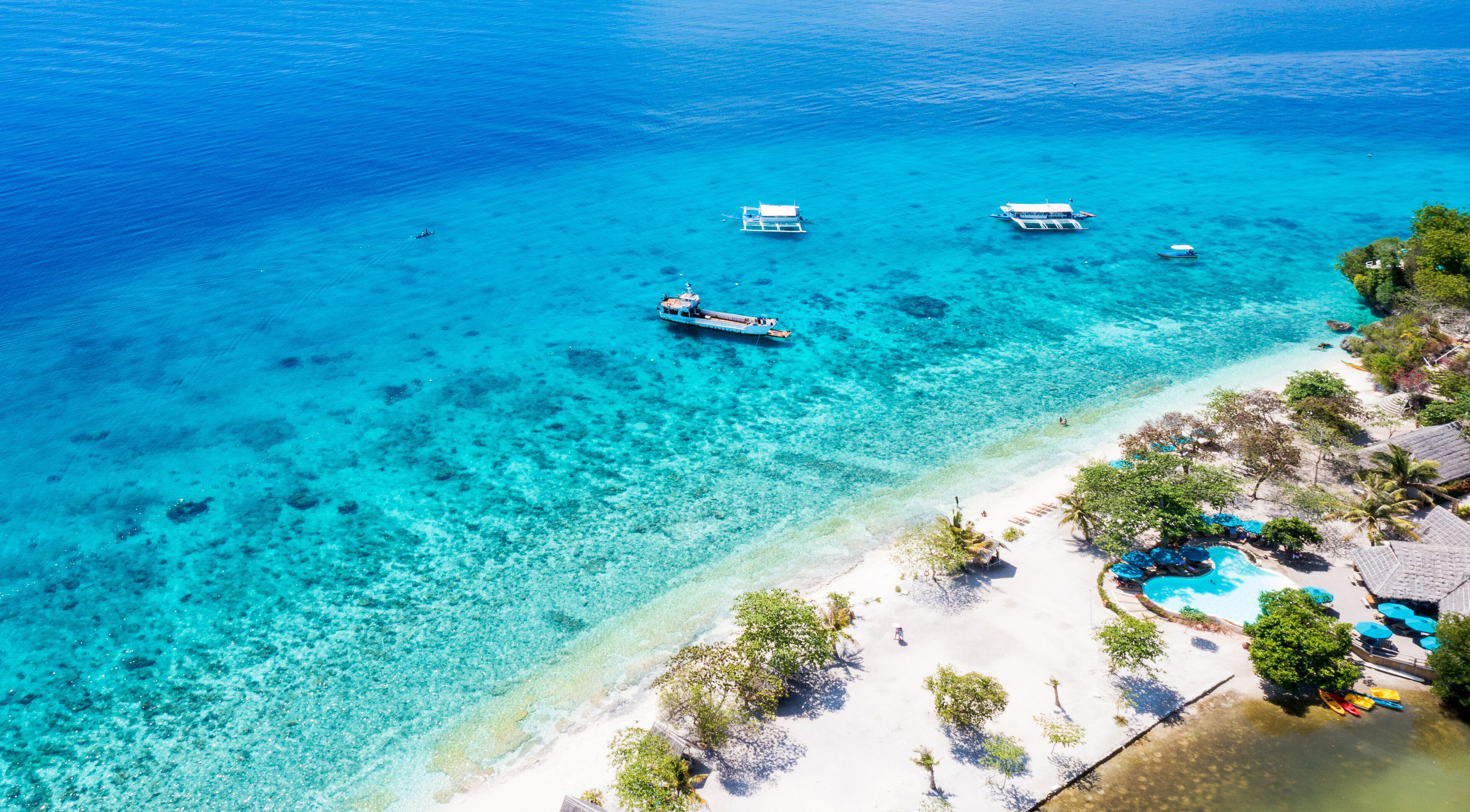 A turquoise sea with boats, a sandy beach, and a pool area with umbrellas.