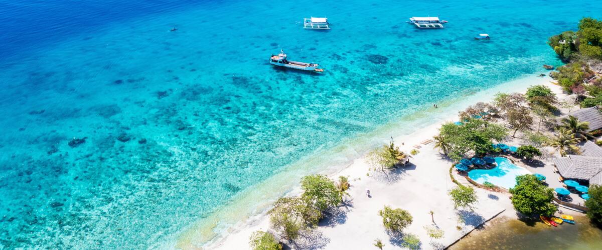 A turquoise sea with boats, a sandy beach, and a pool area with umbrellas.