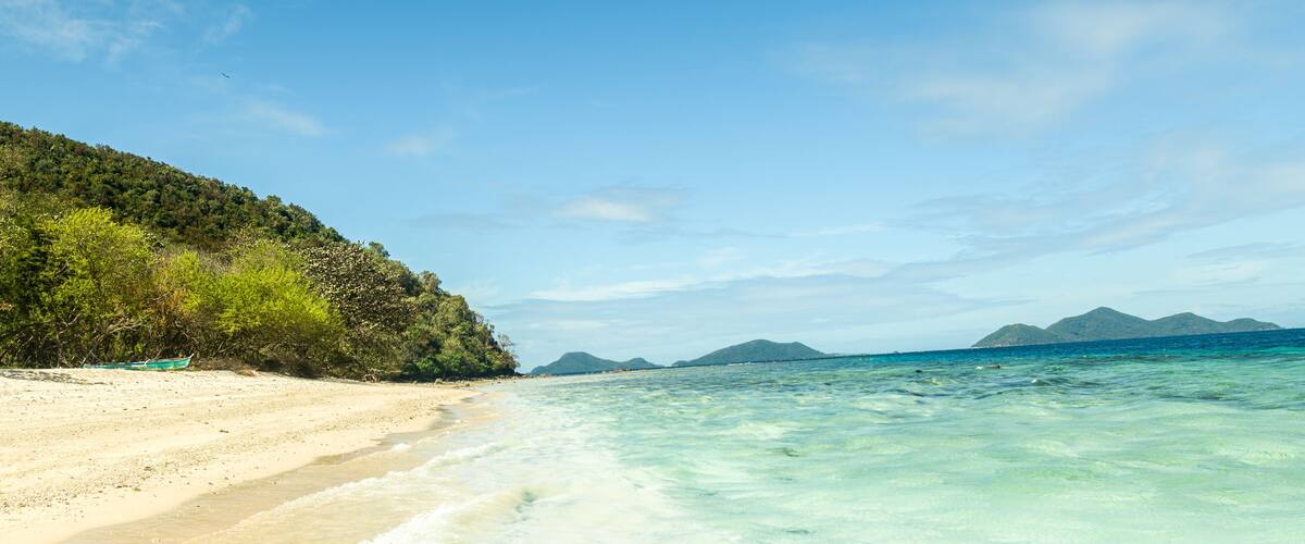 A beach with clear turquoise water, a sandy shore, and a forested hill in the background.
