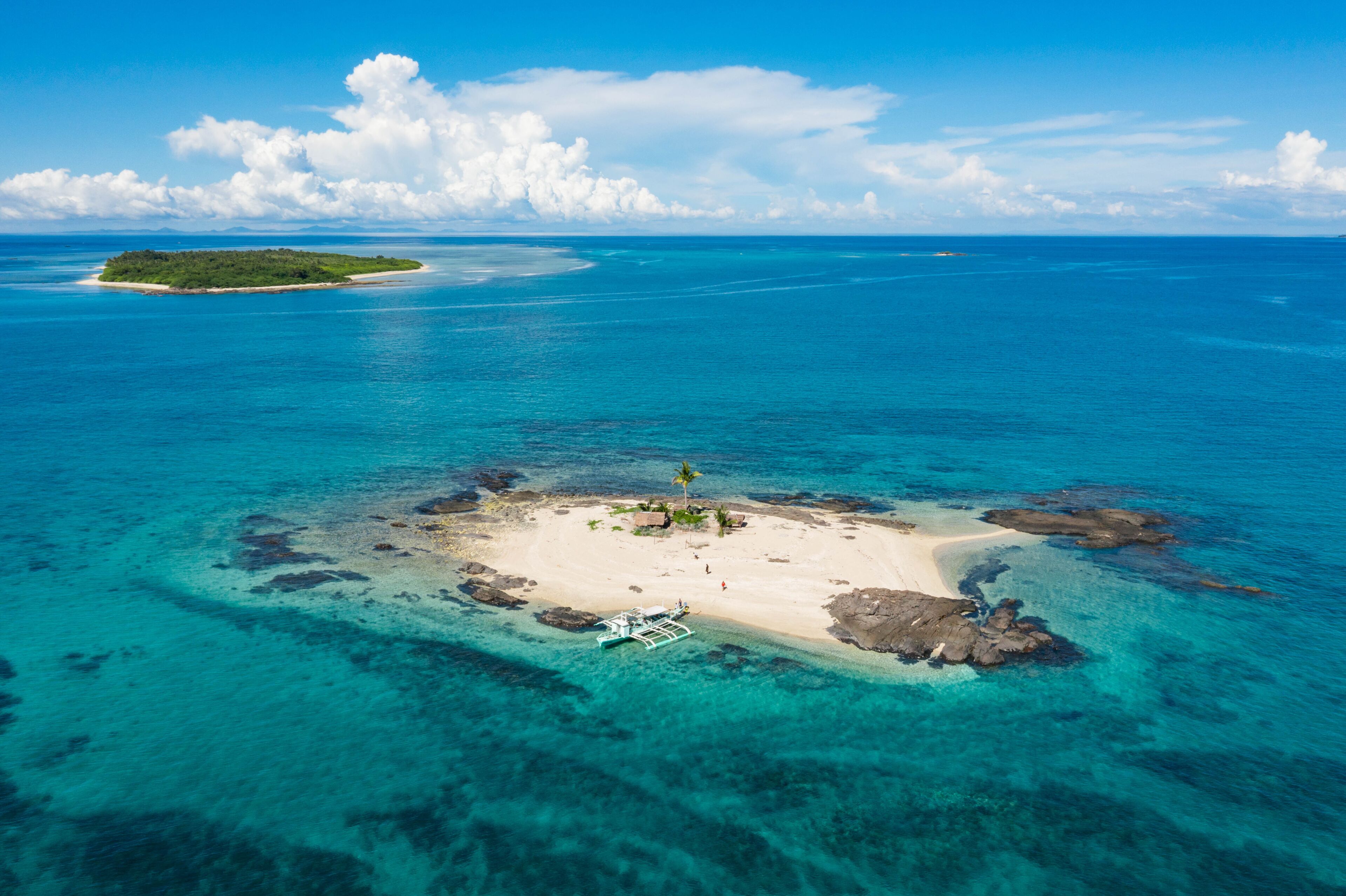 Aerial view of a small sandy island surrounded by turquoise waters, with rocky outcrops and distant lush island, Bancal, Western Visayas, Philippines.