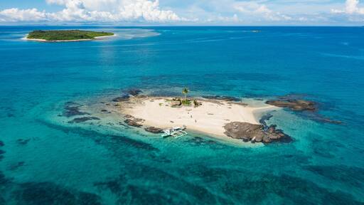 Aerial view of a small sandy island surrounded by turquoise waters, with rocky outcrops and distant lush island, Bancal, Western Visayas, Philippines.
