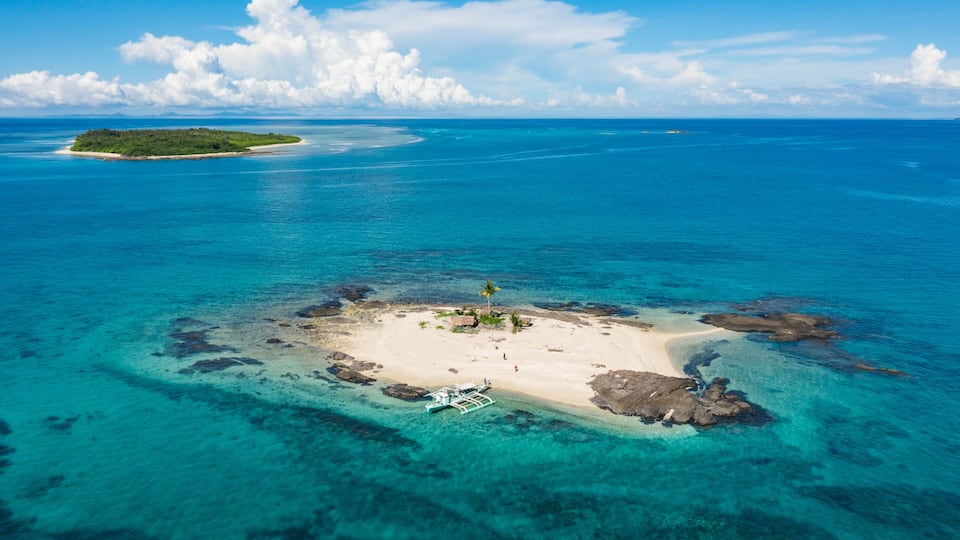 An aerial view of a turquoise island with clear waters, a sandy beach, and a small boat anchored near the shore.