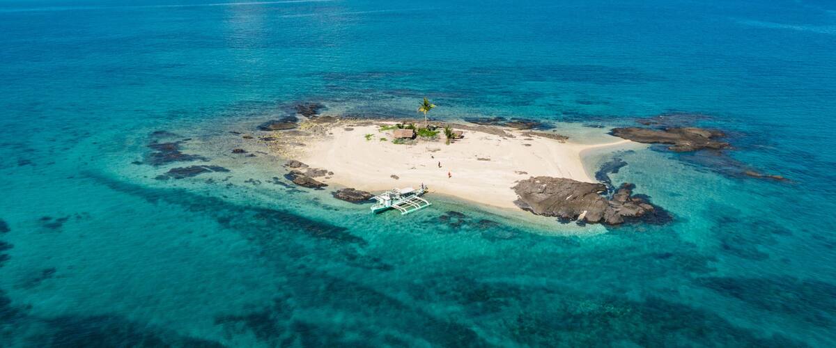An aerial view of a turquoise island with clear waters, a sandy beach, and a small boat anchored near the shore.